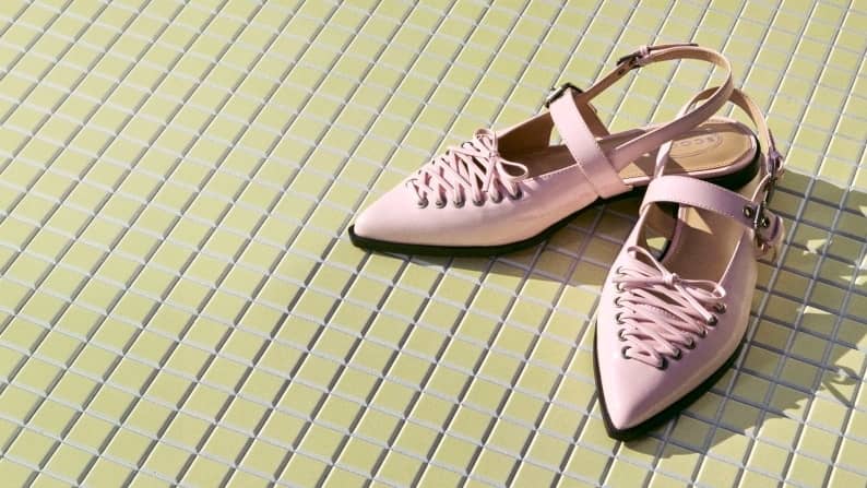 Pair of blush pink pointed toe slingback flats with lace up detailing and low black soles resting on a tiled surface.