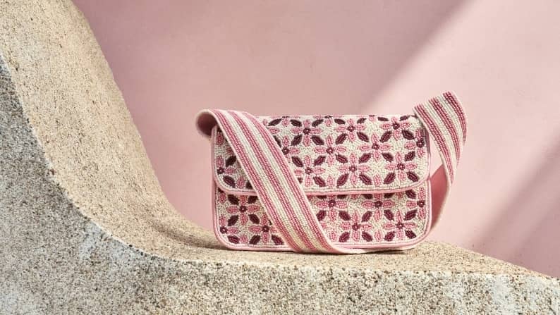 Beaded shoulder bag with pink and cream floral pattern and wide striped strap resting on a stone ledge against a pink wall.