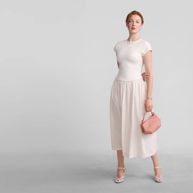 A woman wearing a cream dress and heels is holding a pink bag.