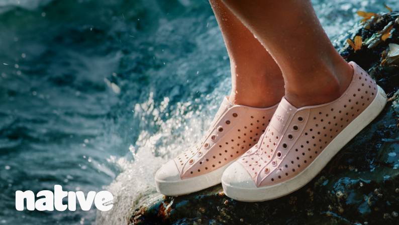 Native logo in the bottom left corner. A woman wearing pink shoes is standing on a rock on the bank of a river.