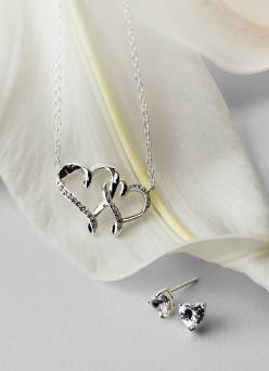 A silver double heart pendant and a pair of silver stud earrings are layered on cream fabric.