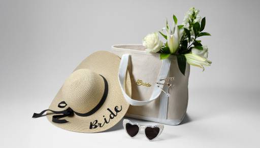 A straw hat with “bride” in black letters, a cream and white tote bag, and a pair of white heart-shaped sunglasses.