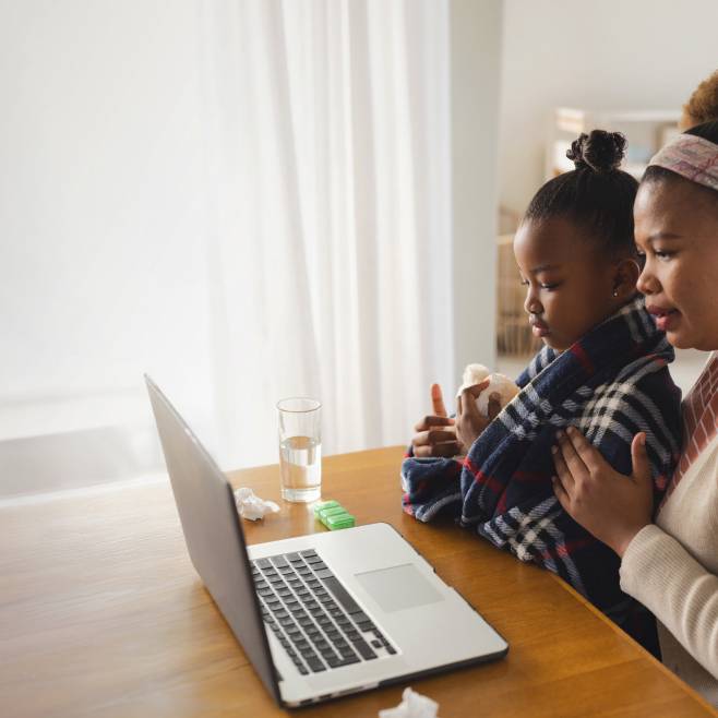 A mother sits beside her child, comforting them while they look at a laptop during a telehealth visit.