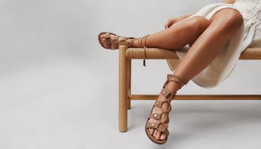 A close-up image of a woman’s legs is shown. She is sitting on a bench and wearing brown gladiator sandals.