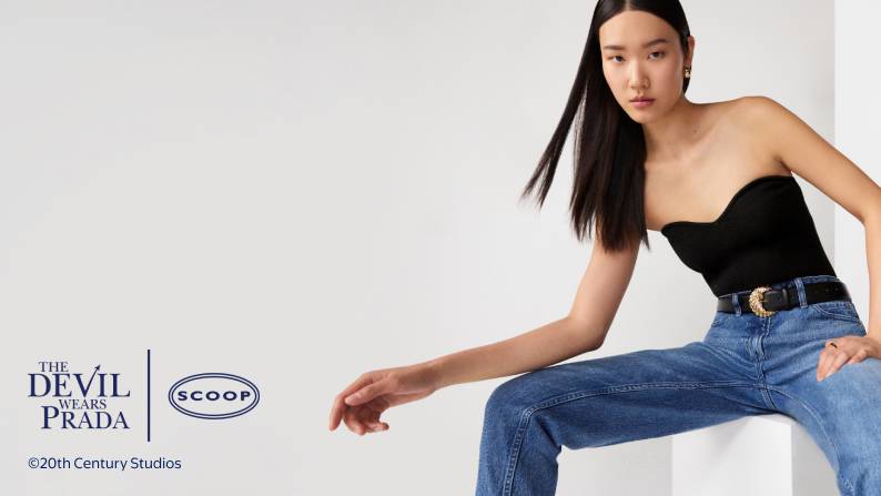 Model sits in studio wearing black strapless top and blue jeans with black belt and gold buckle looking at camera.